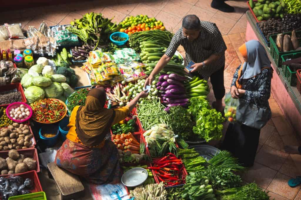 Blick von oben auf zwei Menschen beim Einkauf auf einem Gemüsemarkt mit am Boden sitzender Verkäuferin