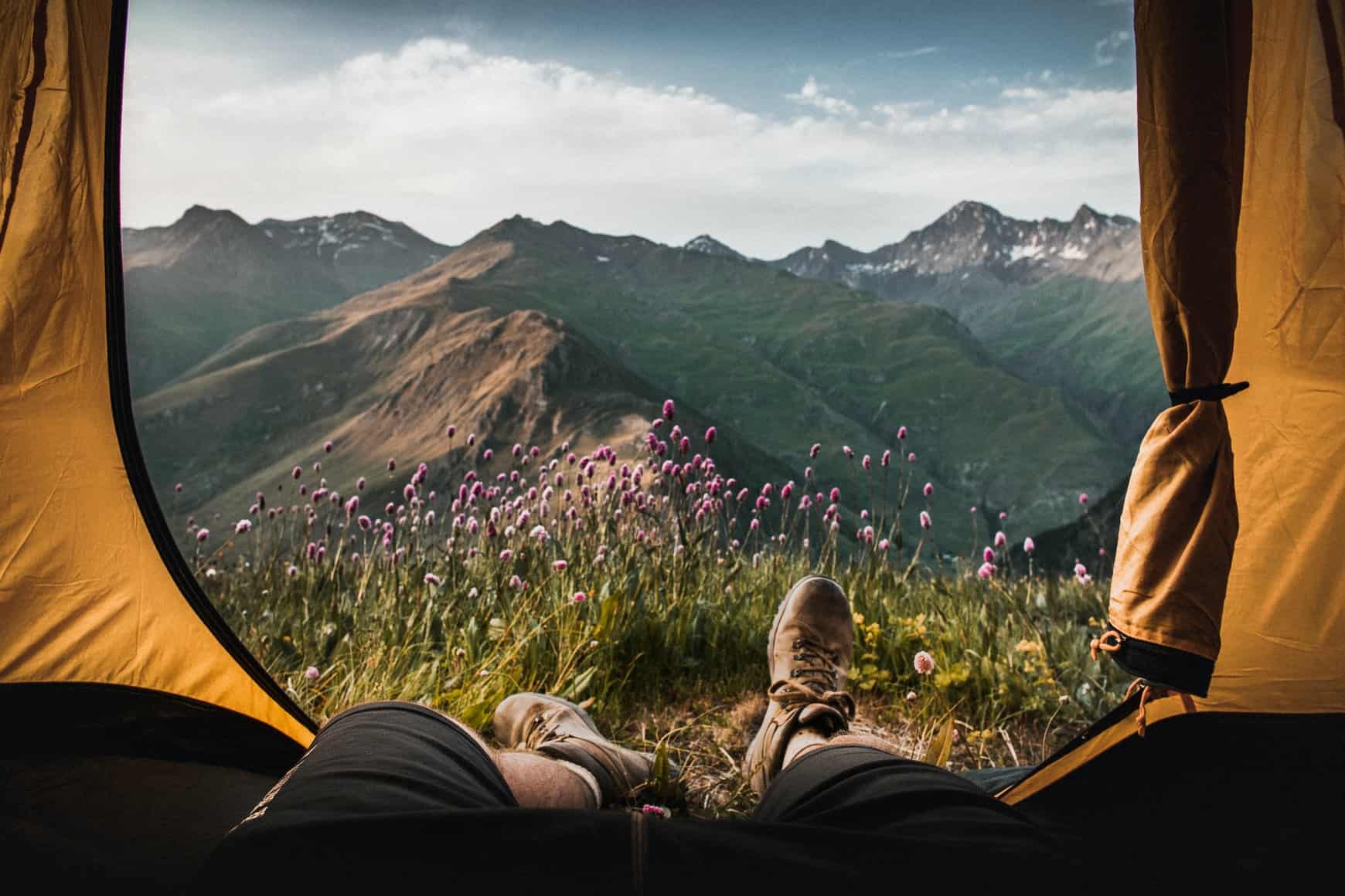 Blick aus dem Zelt auf Bergpanorama mit Beinen und Wanderschuhen im Vordergrund